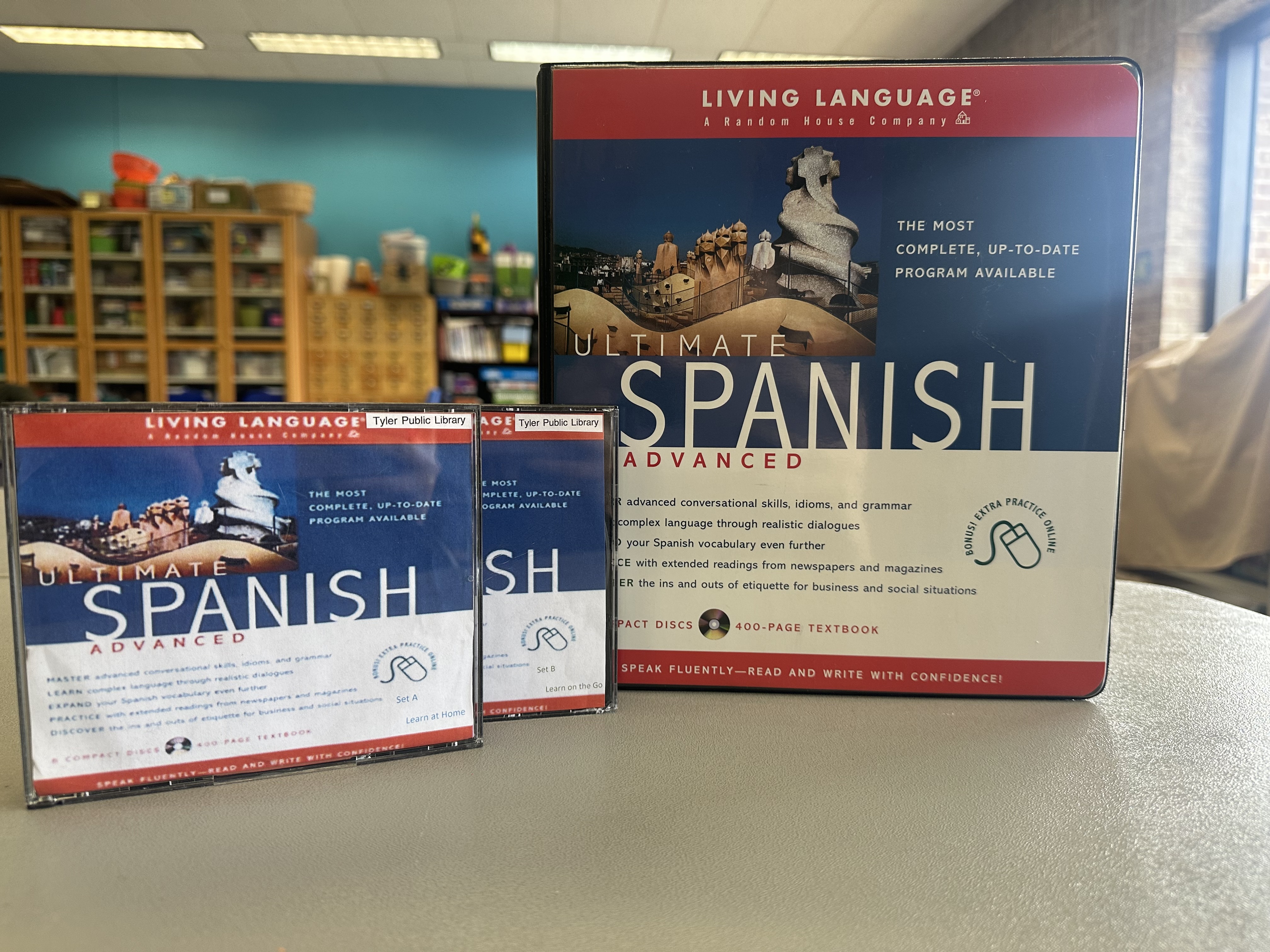 Contents of an Adult Language Kit on a grey table in the Library's Makerspace room. Contents are a Living Language Advanced Spanish course with a book and two CD-ROM cases that are part A and part B. There are blue walls in the background with craft supply shelves and storage. 