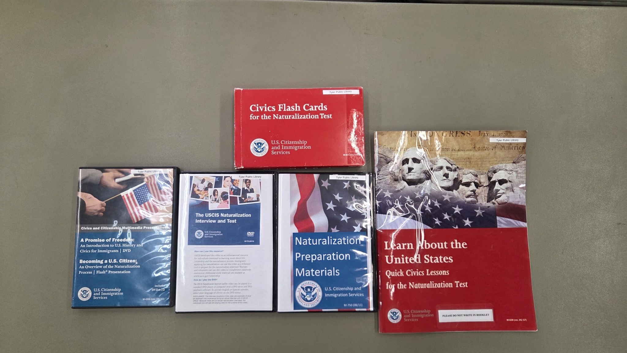 Contents of a Citizenship Kit on grey table. The contents are a set of Civics Flash Cards, three DVDs of Naturalization Interview and Test prep, and one Naturalization study book.