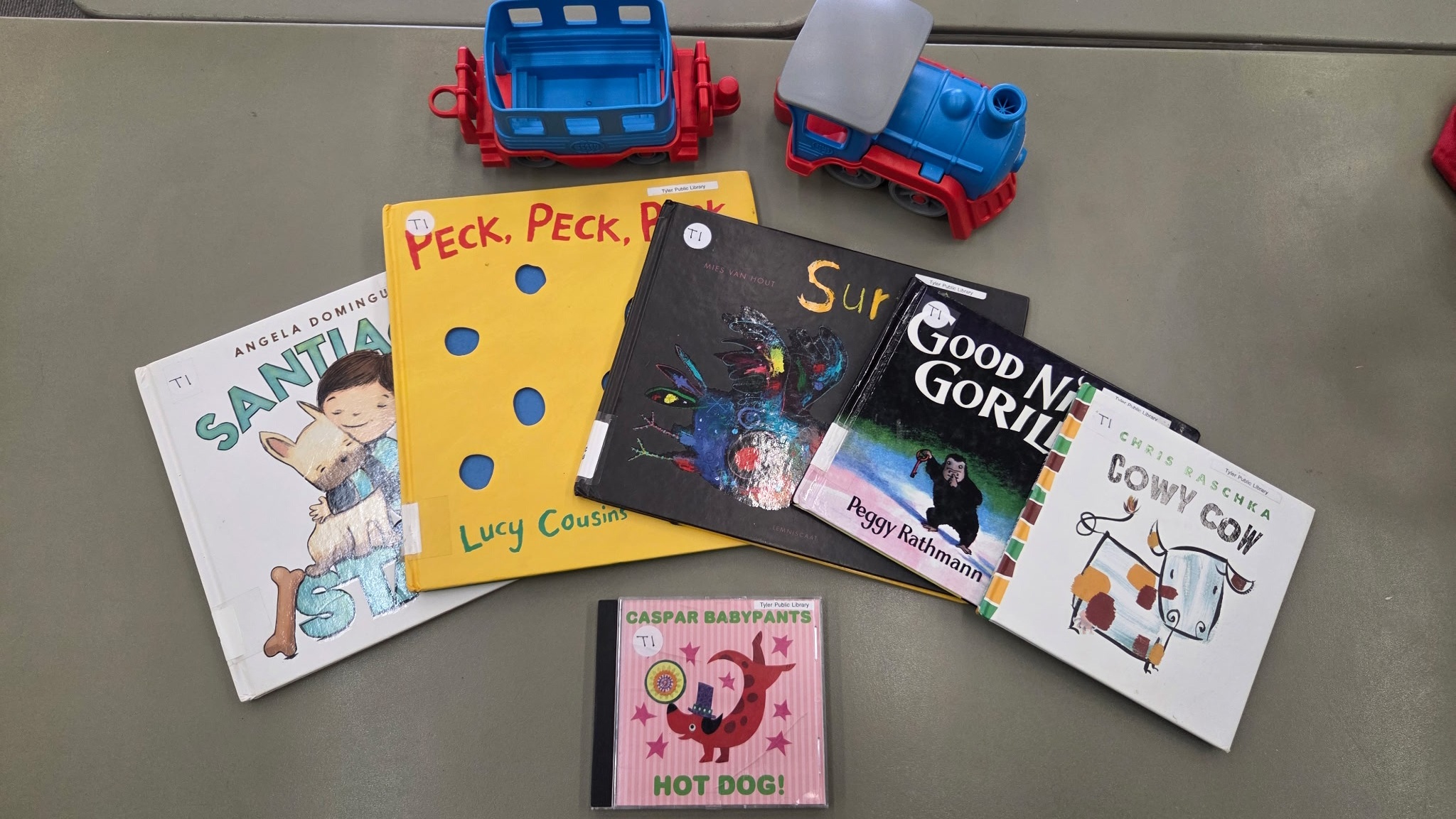 Contents of an Early Literacy Kit on a grey table. The contents are a red and blue toy train, a CD called Caspar Babypants Hot Dog!, and five picture books.