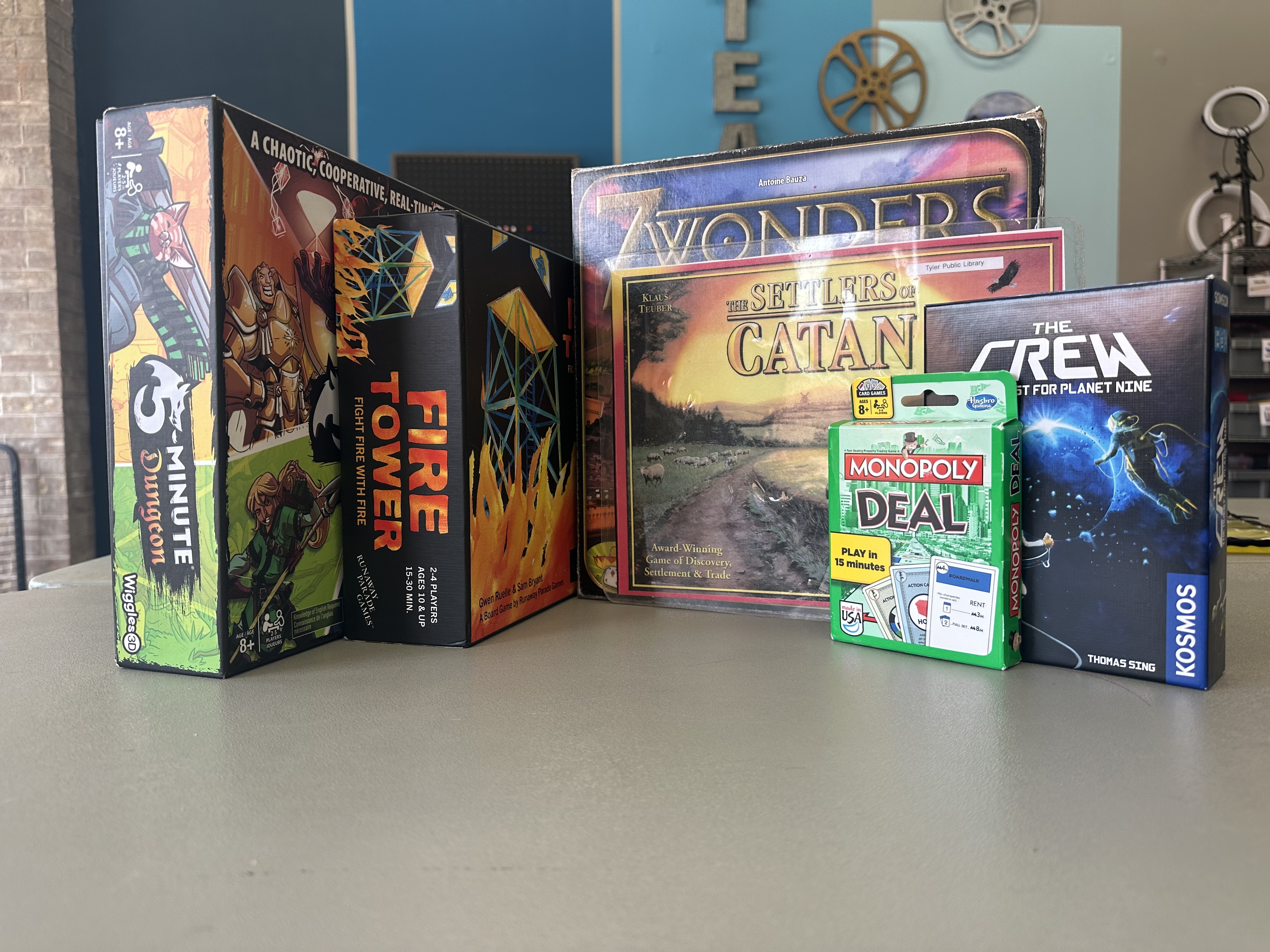 A selection of six board games from the library's collection on a table in the Makerspace room. The games are Five-Minute Dungeon, Fire Tower, Seven Wonders, Settlers of Catan, The Crew, and Monopoly Deal. The background is a blue wall with gears on it.