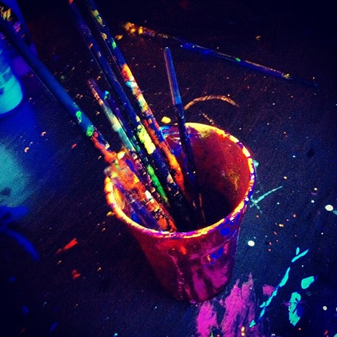 A photo of uncleaned paintbrushes inside a cup on a table with splattered paint. Everything is lit by blacklight.