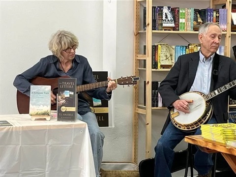 Folk duo, Stringfellows, play in a book shop. Lead guitarist TJ Hassell sits behind a table displaying one of her books, A Fortunate Turn. Her husband, Stuart, sits to the right of her playing a banjo in front of a shelf of various books.